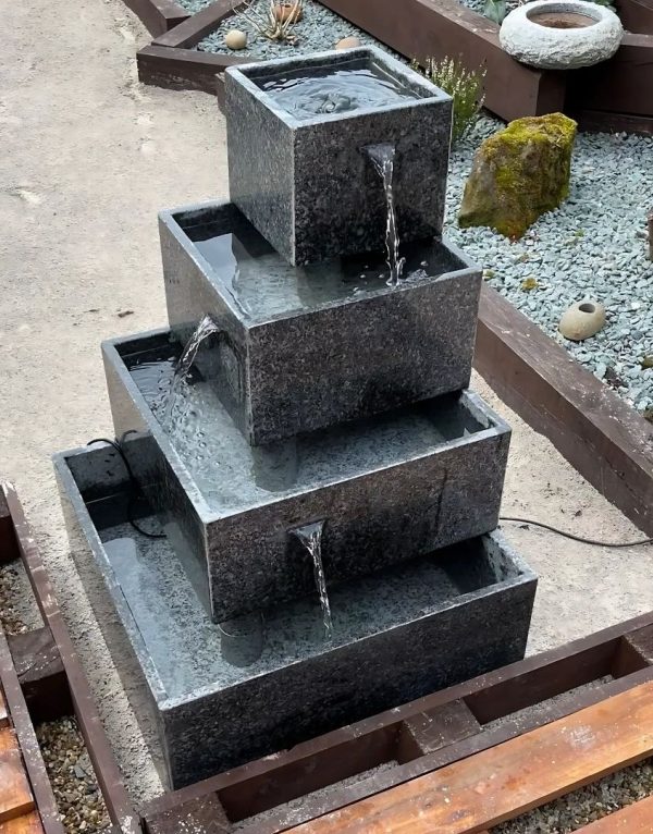 Tiered granite water feature with four square cascading pools, set in a landscaped garden area with gravel, rocks, and wooden borders