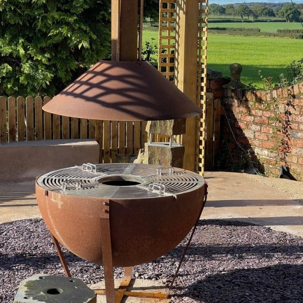 Corten steel 'Timpani Drum' BBQ grill with stainless steel griddle and optional chimney hood, set on a patio with countryside views in the background