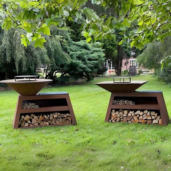 Two corten steel outdoor BBQ grills with wood storage compartments, displayed on a grassy lawn surrounded by trees and a garden setting