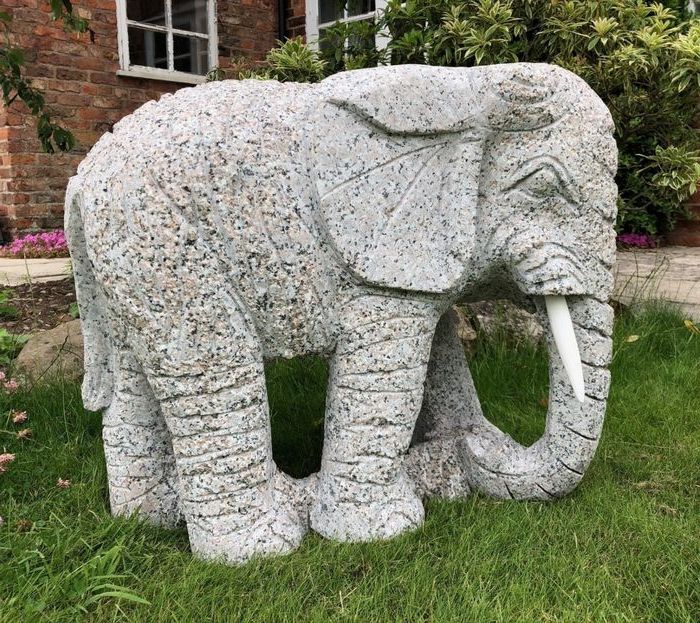 Hand-carved granite elephant statue with white tusks, placed on a grass lawn in front of a brick house and garden shrubs