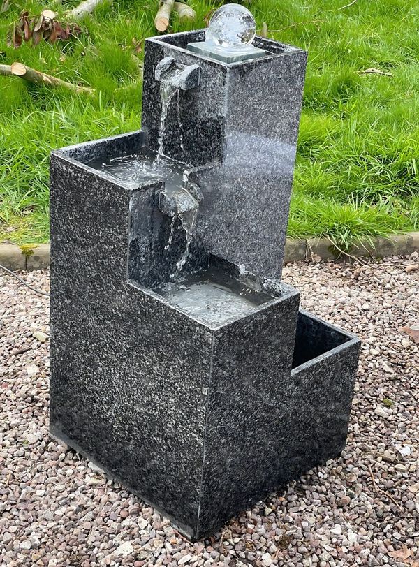 Tiered granite water feature with a glass sphere on top, cascading water into three levels, placed on a gravel surface in a garden setting